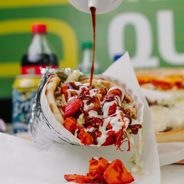 Sauce being drizzled onto a Halal Munchies chicken wrap with white and red sauces, and a soda in the background.