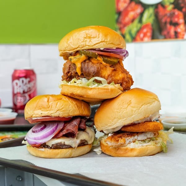A stacked trio of Halal Munchies burgers: fried chicken, beef pastrami, and fish, with a Dr. Pepper can.